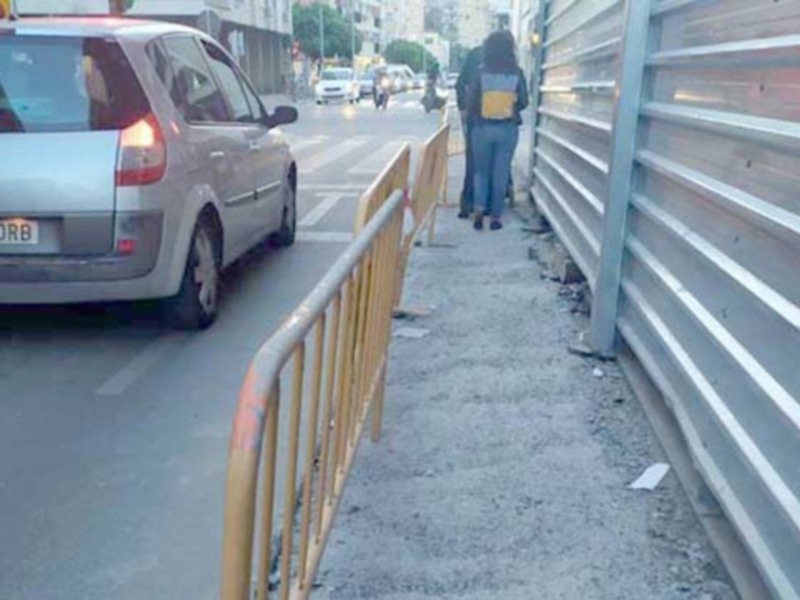Vista de una calle con un coche estacionado y una persona caminando cerca de una cerca metálica.