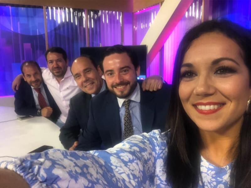 Grupo de personas sonrientes en un estudio de televisión con una mujer en el centro y tres hombres a su izquierda. El fondo es un estudio de TV con luces brillantes y colores vibrantes.