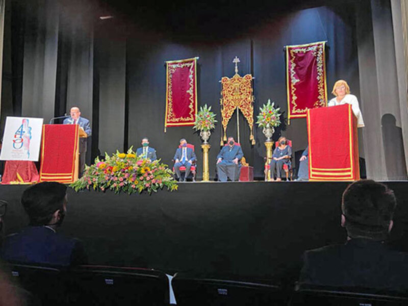 Una imagen de una ceremonia formal con un grupo de personas sentadas en una mesa grande, rodeadas por flores y mantel rojo. En el fondo hay dos tapices con bordados dorados y rojos, una cruz en el centro y un símbolo azul con letras blancas. La escena parece ser parte de una celebración o evento importante, posiblemente religioso.