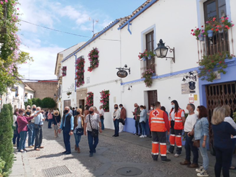 Una multitud de personas camina por una calle estrecha, rodeada de edificios blancos con detalles azules. En la esquina, un grupo de personas espera en fila, mientras que otros caminan hacia adelante. En la esquina opuesta, personal de emergencia con chalecos naranjas espera junto a una farola. Las fachadas están adornadas con flores y plantas, y el cielo es azul con algunas nubes.