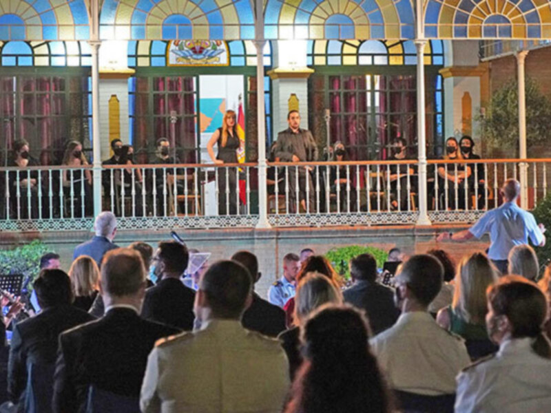 Una imagen de una audiencia en un evento formal, con personas vestidas formalmente. En el fondo, se pueden ver vitrales coloridos y una estructura de madera con un escudo. La audiencia está sentada en filas, mirando hacia adelante.