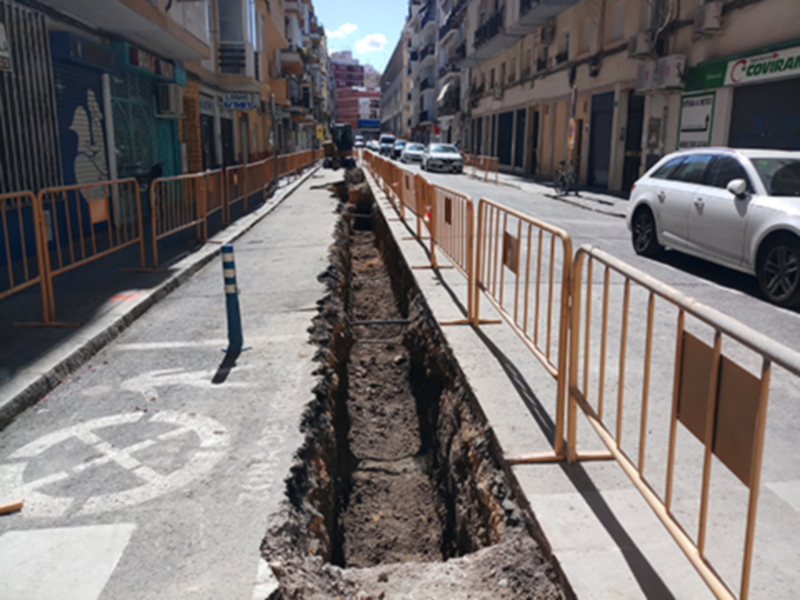 Un túnel de excavación en la calle, con una carretera y edificios al fondo. La imagen muestra un área de construcción o reparación en la calle, con una barrera de protección y una carretera a un lado.
