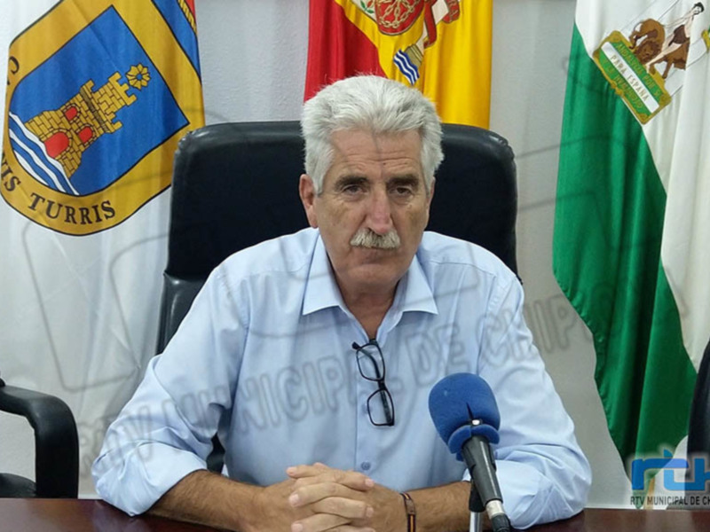 Persona sentada en una mesa con un micrófono, ante banderas y escudos de armas, probablemente en una conferencia de prensa o reunión oficial.