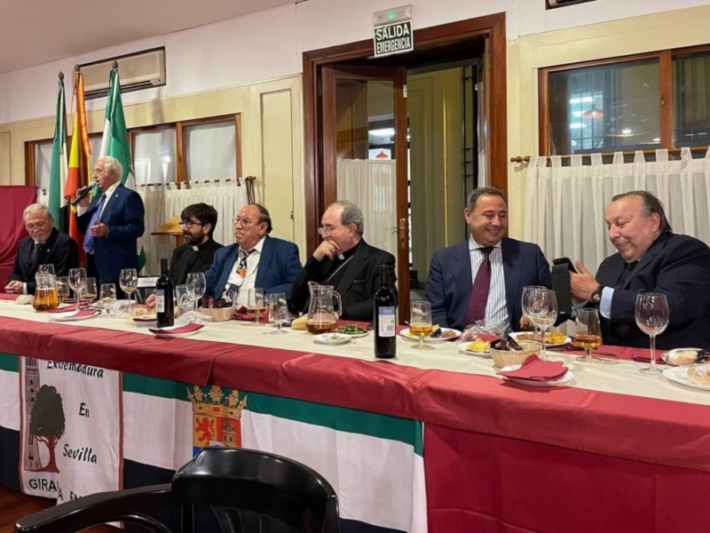 Una reunión formal con siete hombres sentados en una mesa cubierta de un mantel rojo y blanco, con banderas de varios países en el fondo. La mesa está servida con copas y botellas, y hay un teléfono móvil en la mesa. La imagen se toma en una sala con puertas abiertas y ventanas.