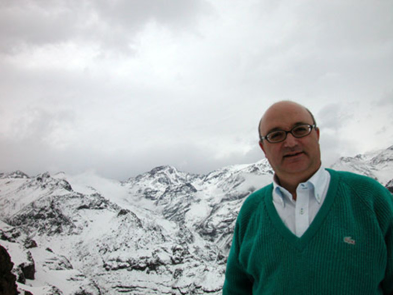 Un hombre con una chaqueta verde y una camisa blanca posa frente a un paisaje nevado con montañas en el fondo. El cielo está cubierto de nubes grises y blancas, creando un ambiente tranquilo y frío.