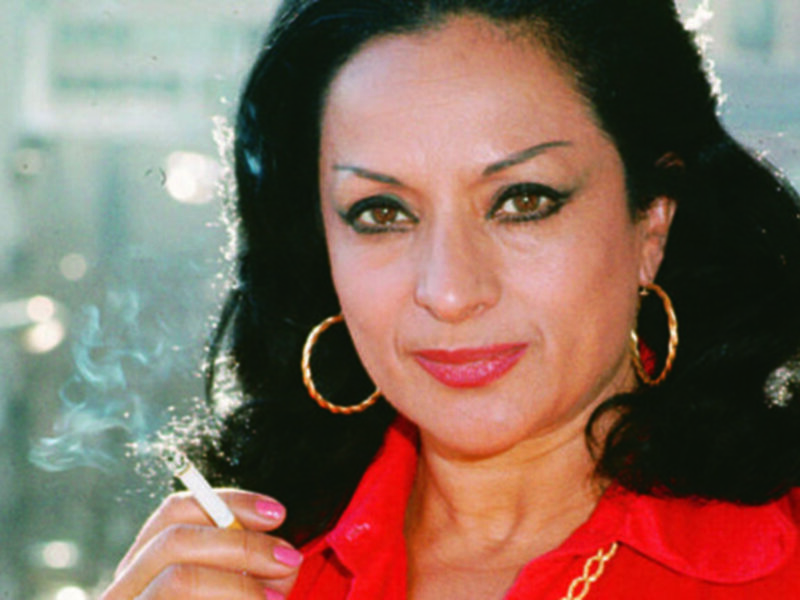 A una mujer con cabello oscuro y ondulado, usando un chaleco rojo y joyas doradas. Está fumando una cigarrillo, con un fondo borroso que parece ser una ventana o vidrio.