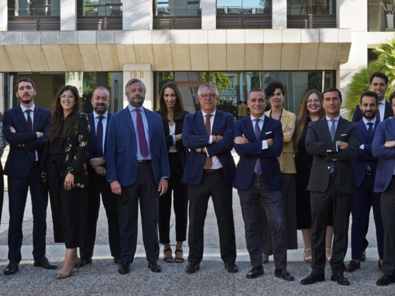 Una imagen de una fila de personas vestidas con trajes y chaquetas, posando frente a un edificio moderno. La escena está iluminada por el sol, con palmeras y árboles al fondo.