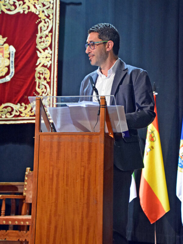 Hombre hablando en un podio con un fondo de bandera española y un escudo heráldico.