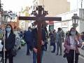 Personas con máscaras faciales y cruces durante una procesión religiosa en una calle con edificios y gente al fondo.
