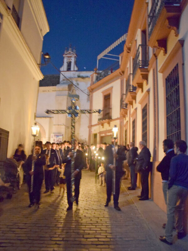 Procesión nocturna con cruces y velas en una calle adoquinada de Sevilla.