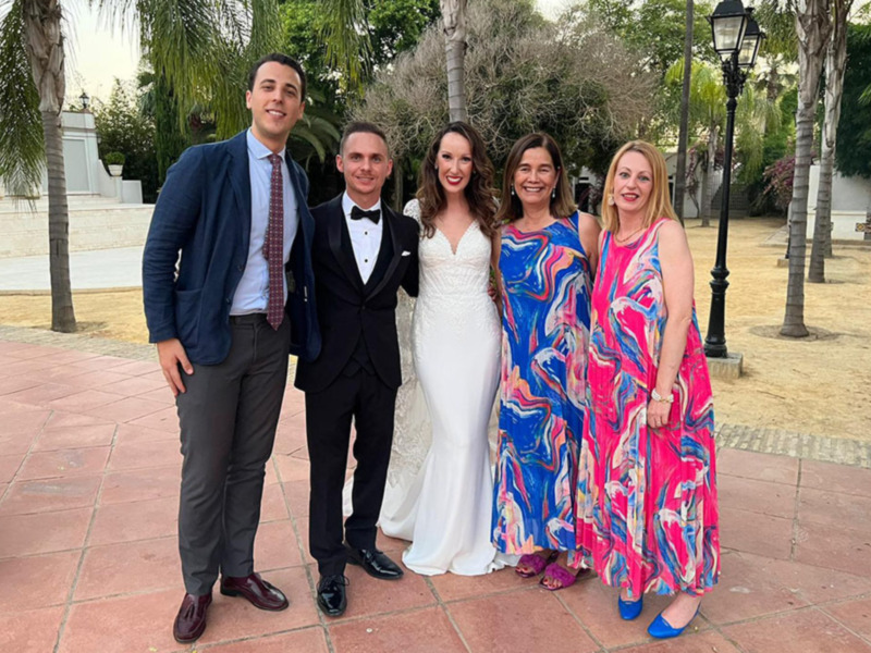 Una familia posa juntos en un evento formal, con un hombre en traje y dos mujeres en vestidos coloridos, frente a un parque con palmeras y una lámpara de calle.