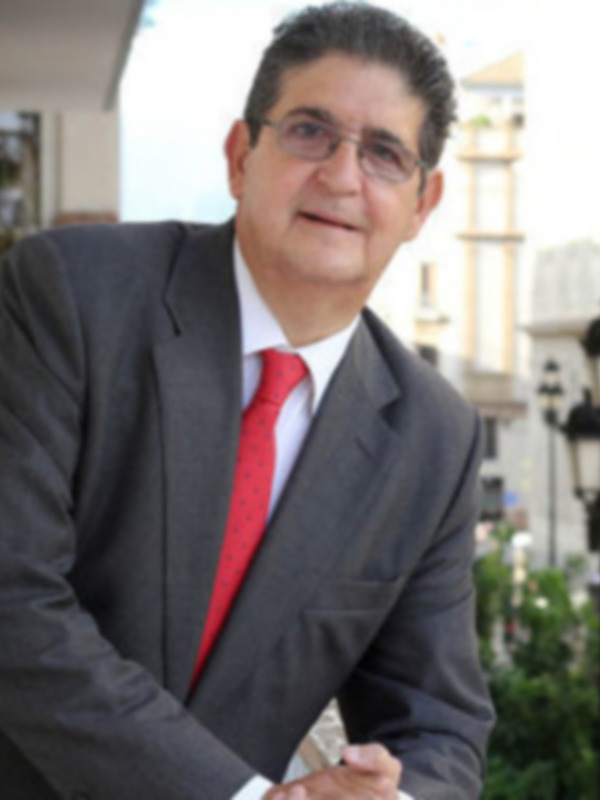 Un hombre en traje y corbata roja, posando para la foto en un entorno urbano con edificios y farolas al fondo.