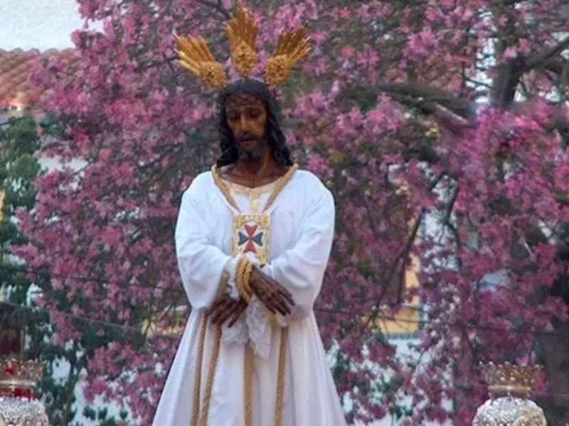 Un hombre vestido con una túnica blanca y dorada, posiblemente representando a Jesús, se encuentra en un entorno con árboles frondosos y flores rosadas. La escena parece ser parte de una procesión religiosa o ceremonia tradicional, con detalles que sugieren un contexto histórico o cultural específico.