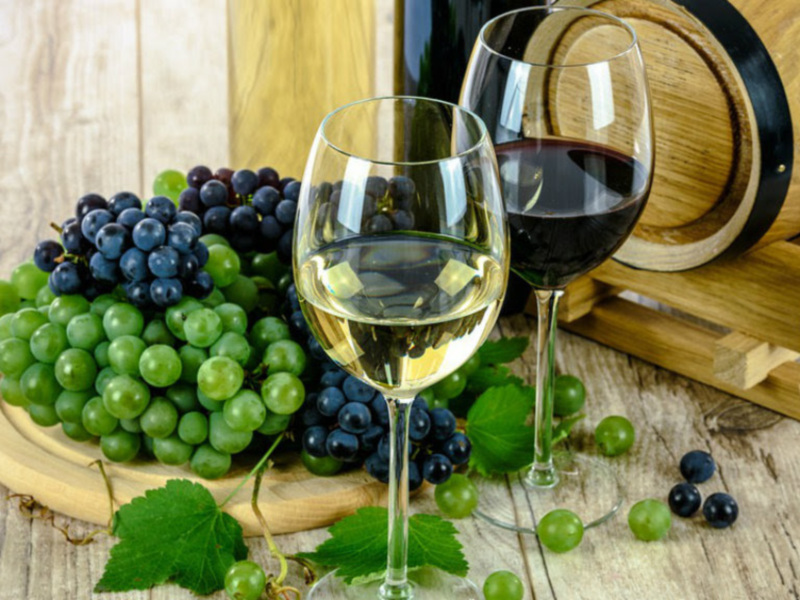 Vino blanco y tinto en copas, uvas verdes y negras frescas, barril de madera al lado.