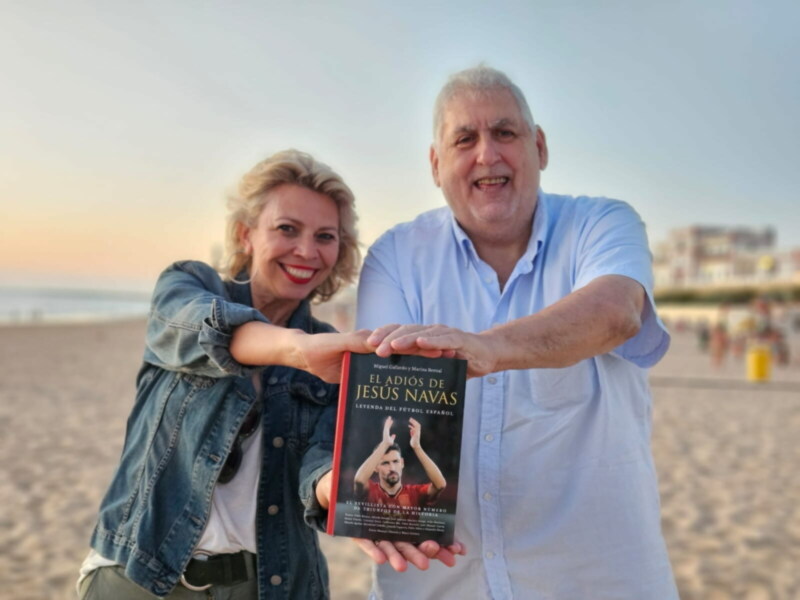 Autor y editor presentando 'El Adiós de Jesús Navas', libro sobre el futbolista español, en la playa al atardecer.