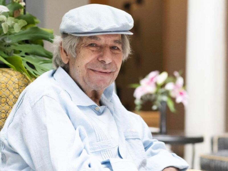Un hombre con una gorra azul sentado en un sillón de terciopelo amarillo, con flores y una puerta detrás.
