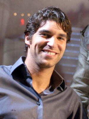 Un hombre con cabello oscuro y ondulado, sonriendo ampliamente. Está vestido con una camisa de manga larga gris y un chaleco negro, posiblemente en un evento o conferencia. El fondo es borroso pero parece ser una sala con luces y estructuras metálicas.