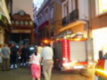Un camión de bomberos está estacionado en la calle, con una persona vestida de blanco caminando hacia él. La imagen muestra un entorno urbano con edificios históricos y gente observando la escena.