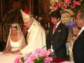 Un matrimonio recién casado, el novio y la novia, firman un documento en una ceremonia religiosa. El padre de la novia y otros invitados observan el momento. La imagen se realiza en un entorno decorado con flores y madera, probablemente en una iglesia.