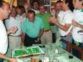 Celebración con amigos en un ambiente festivo, donde se disfruta de una torta decorada con flores verdes y blancas. La celebración parece ser en un ambiente informal, con una mesa llena de comida y bebidas. El ambiente es animado y lleno de alegría.