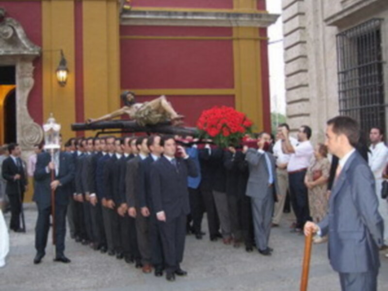 Procesión con imágenes religiosas y flores rojas, en un entorno urbano con edificios históricos.