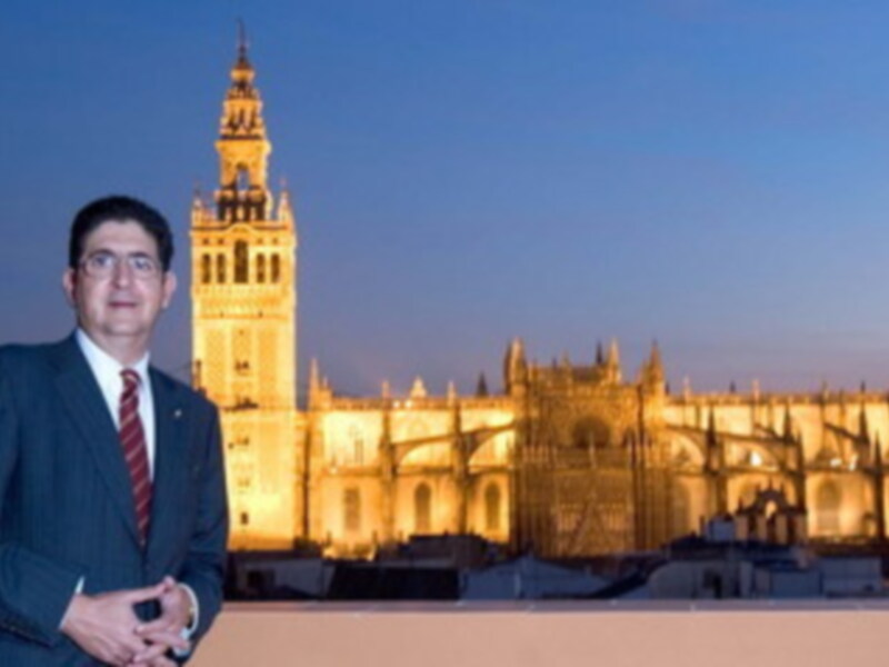 Un hombre en traje y corbata posa frente a la Catedral de Sevilla iluminada por el atardecer, con su famosa torre Giralda en segundo plano.