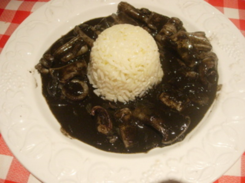 Un plato blanco con un arroz blanco en el centro, rodeado por una salsa negra y varios ingredientes que parecen ser carnes. El plato está sobre un fondo de mesa con una manta roja y blanca.