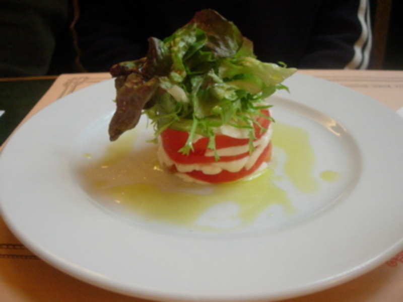 Un delicioso ensalada de tomate con rodajas finas, cubierta por una salsa verde y adornada con hojas de lechuga fresca. El plato se presenta sobre un fondo blanco, destacando la textura y color de los ingredientes.