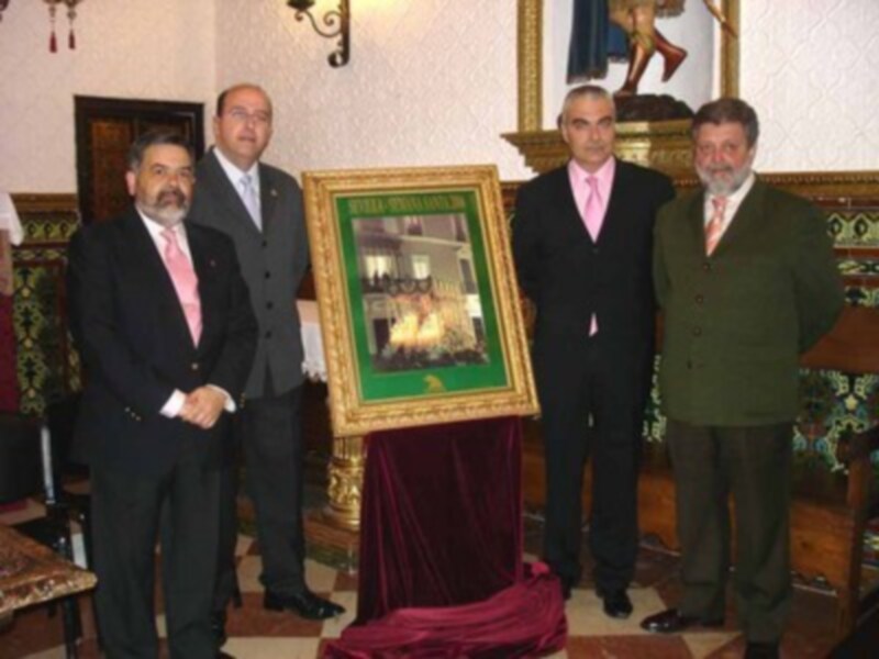 Un grupo de personas en trajes formales posan junto a una imagen dorada y verde, probablemente en un evento formal o ceremonial.