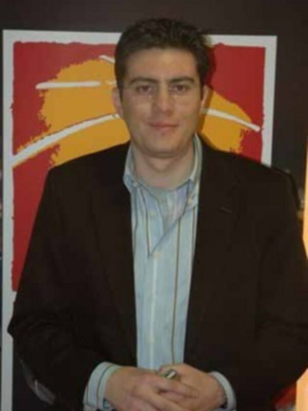 Un hombre con una camisa azul y una chaqueta negra, posando frente a una pared con un arte abstracto rojo y amarillo.