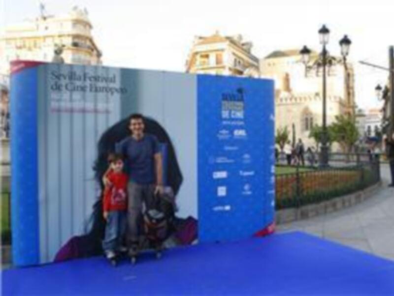 Un hombre y una niña posan en frente de un gran cartel promocional del Festival de Cine Europeo de Sevilla. El cartel es azul con imágenes y texto blanco, y se encuentra en una calle con edificios históricos al fondo.