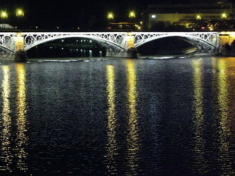 Puente de Triana en la noche, iluminado con luces amarillas reflejándose en el río.