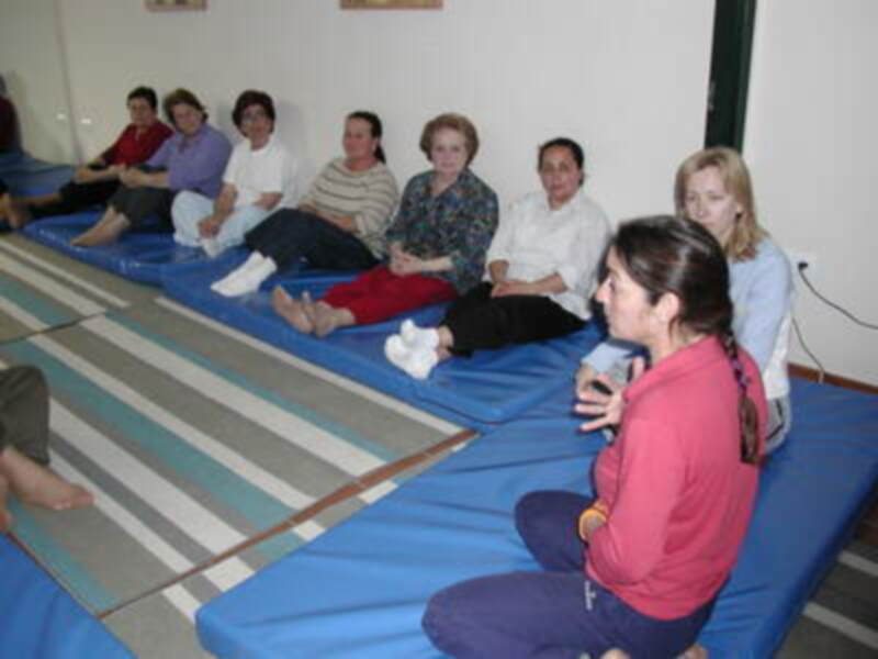 Una grupo de personas sentadas en colchones azules y grises con una cama de colores a rayas. Algunas personas están bebiendo agua.