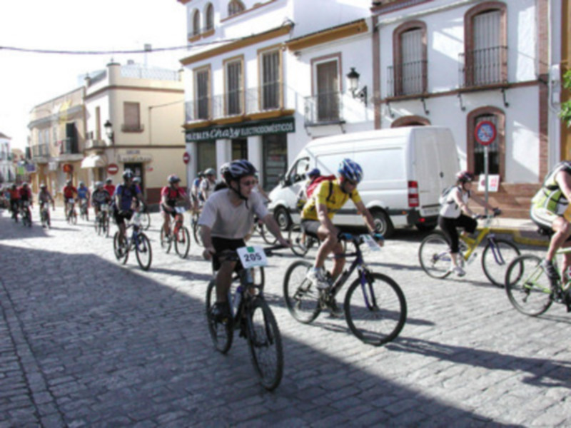 Ciclismo en una calle adoquinada con edificios blancos y azules. Personas en bicicleta, algunos con cascos, en una carrera o actividad recreativa. Suelo adoquinado y edificios con balcones.
