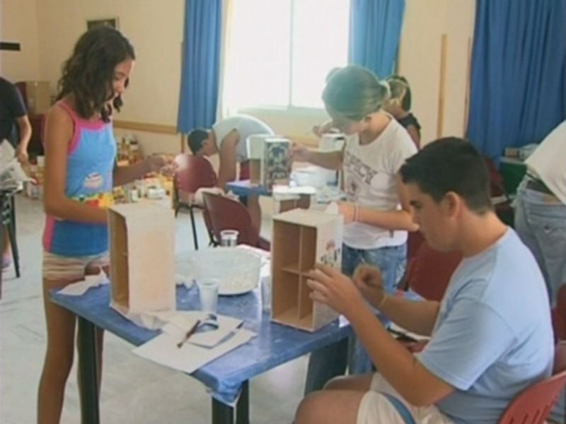 Grupos de niños y adultos trabajando en proyectos artísticos, con paletas y pinturas en la mesa.