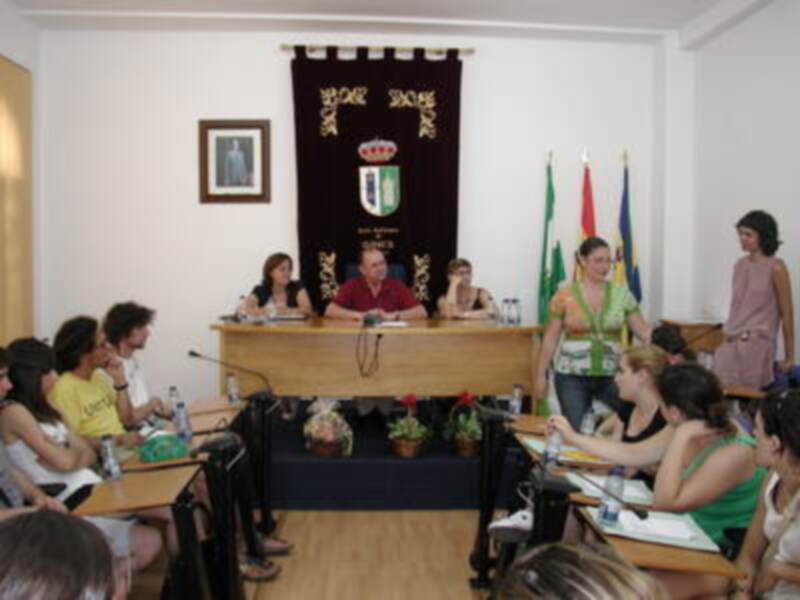 Una sala de conferencias con una mesa central donde se encuentran varias personas sentadas, entre ellas un hombre en el centro y mujeres a su alrededor. En la pared hay una bandera con un escudo y un retrato de una persona enmarcado. El ambiente es formal, probablemente durante una reunión o conferencia.