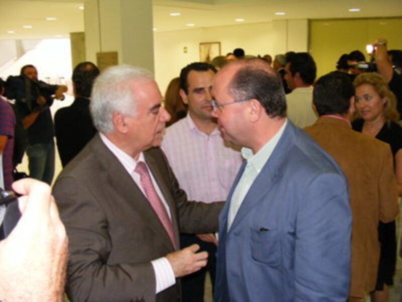 Dos hombres en trajes charlan en un evento, uno de ellos con una camisa blanca y el otro con una camisa azul. El fondo muestra a otros asistentes y una pared de vidrio.