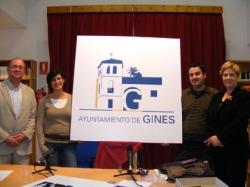 En una sala con cortinas rojas, un grupo de personas sostiene un póster blanco con el logo del Ayuntamiento de Gines y la frase "Ayuntamiento de Gines".
