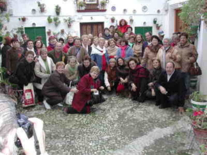 Una multitud de personas posa para una foto en un entorno rural con una casa tradicional al fondo. La imagen muestra a varias personas, tanto jóvenes como mayores, vestidas de manera casual y con sombreros tradicionales. La escena se desarrolla en un camino de piedra, con plantas y flores decorando la casa.