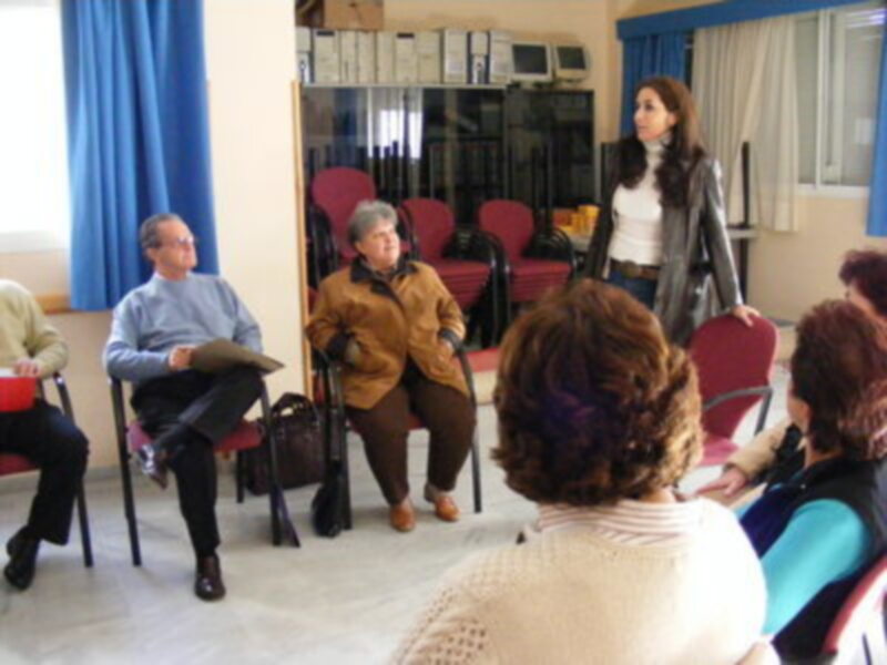 Una persona vestida con una chaqueta marrón y pantalones negros está en pie, mientras otras personas están sentadas en sillas rojas. La imagen parece ser de un evento o reunión formal, con cortinas azules y una pared en el fondo.