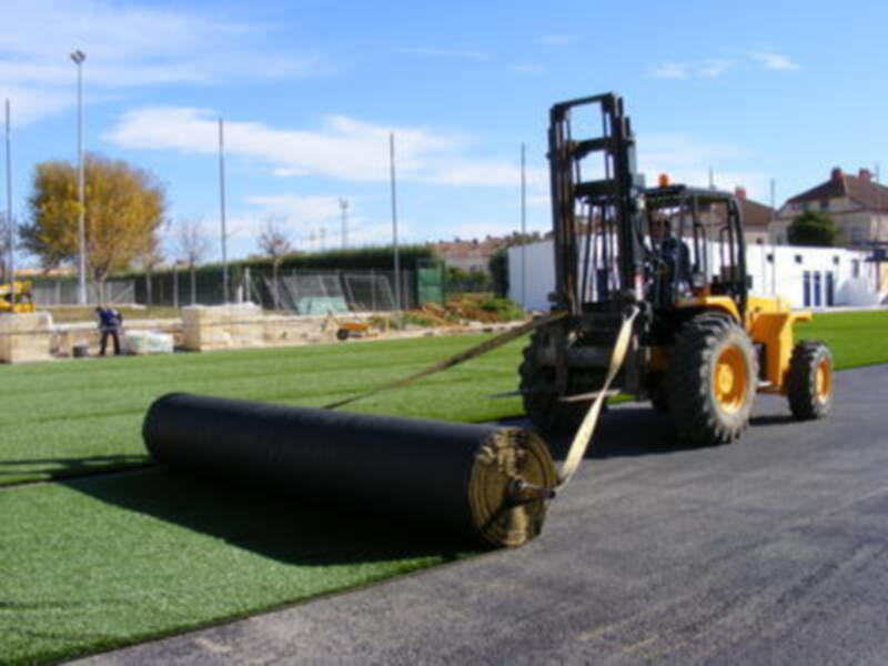 Una máquina de construcción amarilla está colocando un rollo grande de asfalto en una superficie de césped artificial. En el fondo, se pueden ver edificios y árboles con hojas amarillas.