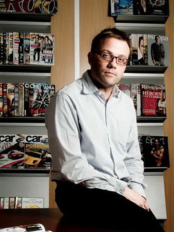 Un hombre con cabello oscuro y gafas se sienta en una mesa frente a un estante lleno de revistas y libros. La imagen muestra una variedad de títulos, incluyendo "Carrozuela" y "Heroes". La escena sugiere un ambiente de trabajo o estudio.