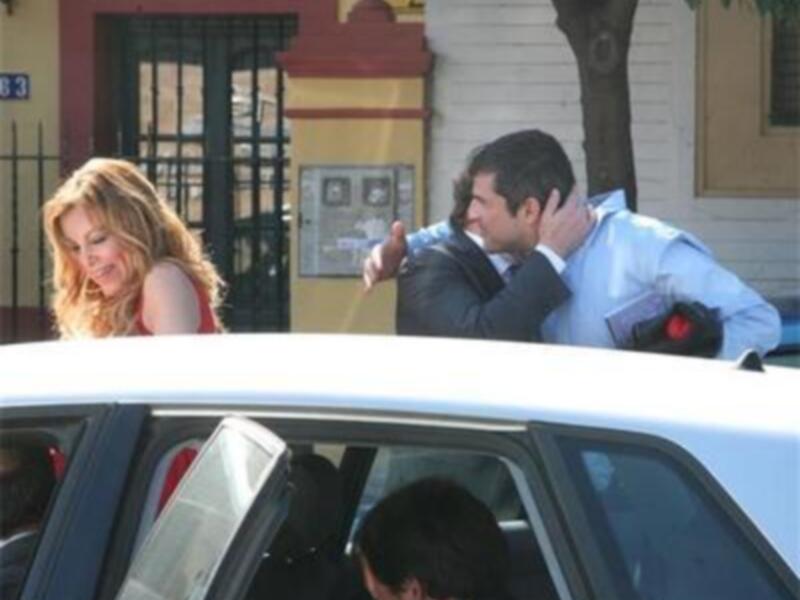 Una mujer con el cabello largo y rubio está hablando con un hombre que parece estar enojado. El hombre tiene la mano en su mejilla y está cerca de un coche blanco. La imagen parece estar tomada en una calle con edificios y un cartel visible al fondo.