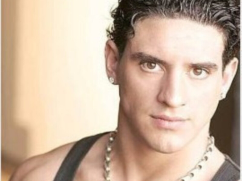 Un hombre con cabello oscuro y rizado, lleva una camiseta negra sin mangas y un collar de metal. El fondo es suave y neutro, enfocándose en el hombre como el protagonista de la imagen.
