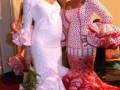 Dos mujeres vestidas con trajes de flamenca posan frente a una pared blanca. La imagen muestra detalles como las mangas de volantes y los vestidos elaborados, típicos del baile flamenco.