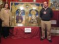 Dos personas posan junto a un cuadro de arte religioso en una exposición. El cuadro muestra dos imágenes de la Virgen y el Niño, rodeadas por ornamentos dorados. La exposición está decorada con mantas rojas y carteles informativos en el fondo.