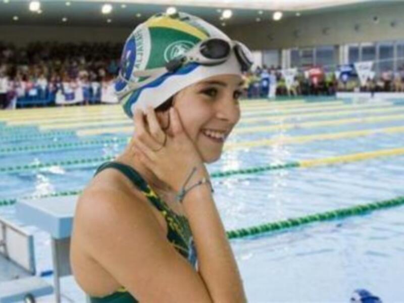 Una joven nadadora sonríe mientras se prepara para competir en una piscina olímpica. La imagen captura el entusiasmo y la concentración antes de una competencia deportiva.