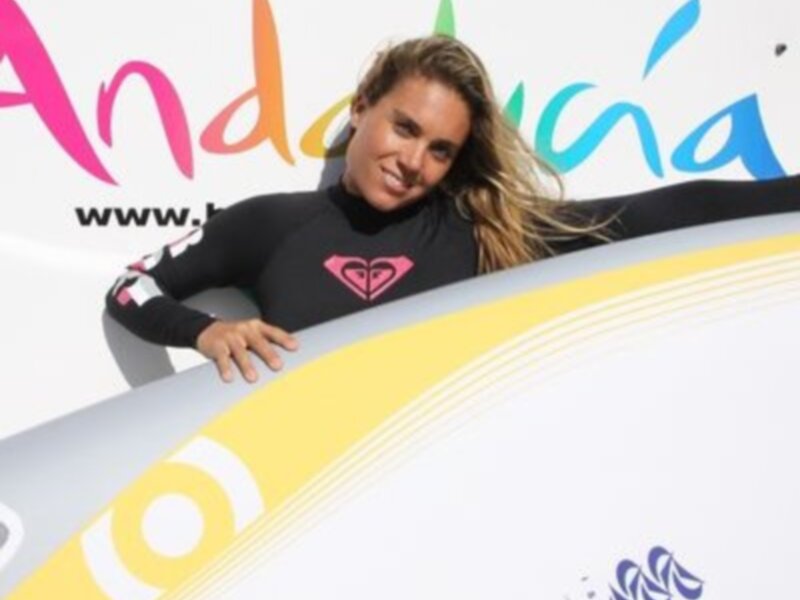 Una persona en traje de neopreno con una camiseta Roxy, posando con una tabla de surf en un contexto de publicidad de Andalucía.
