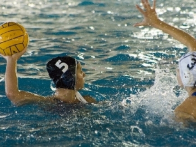 Un jugador de waterpolo con una gorra negra y un n° 5 en la cabeza lanza el balón amarillo hacia otro jugador que se prepara para recibirlo. La acción se desarrolla en un entorno de agua azul claro, con ondas y reflejos que dan vida al juego.