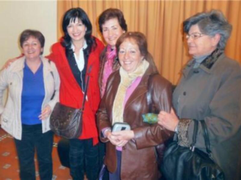 Las mujeres están sonriendo y posando para una foto en un entorno interior con cortinas de fondo. Una de ellas sostiene un teléfono móvil.